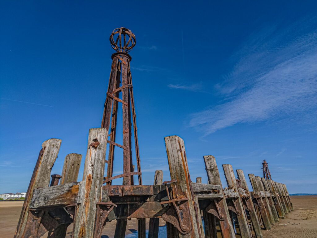 The rusted ironwork and timber piles of St Annes old pier standing on the beach under a clear blue sky.